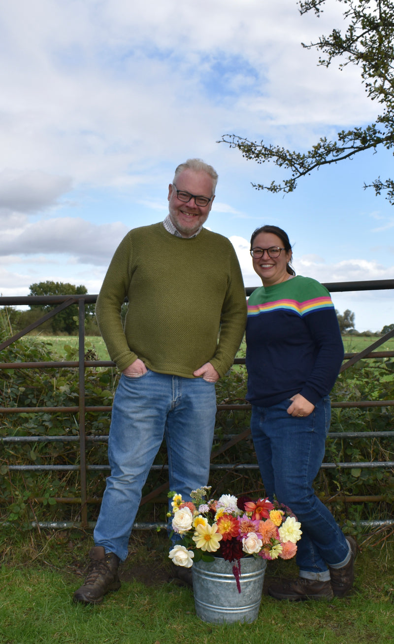 Two people standing outdoors with a metal bucket of flowers, against a backdrop of a metal gate and greenery.