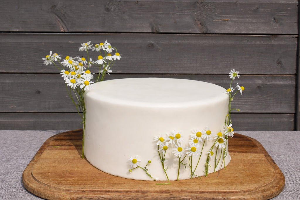 White cake with edible chamomile flowers on a wooden board against a wooden wall.
