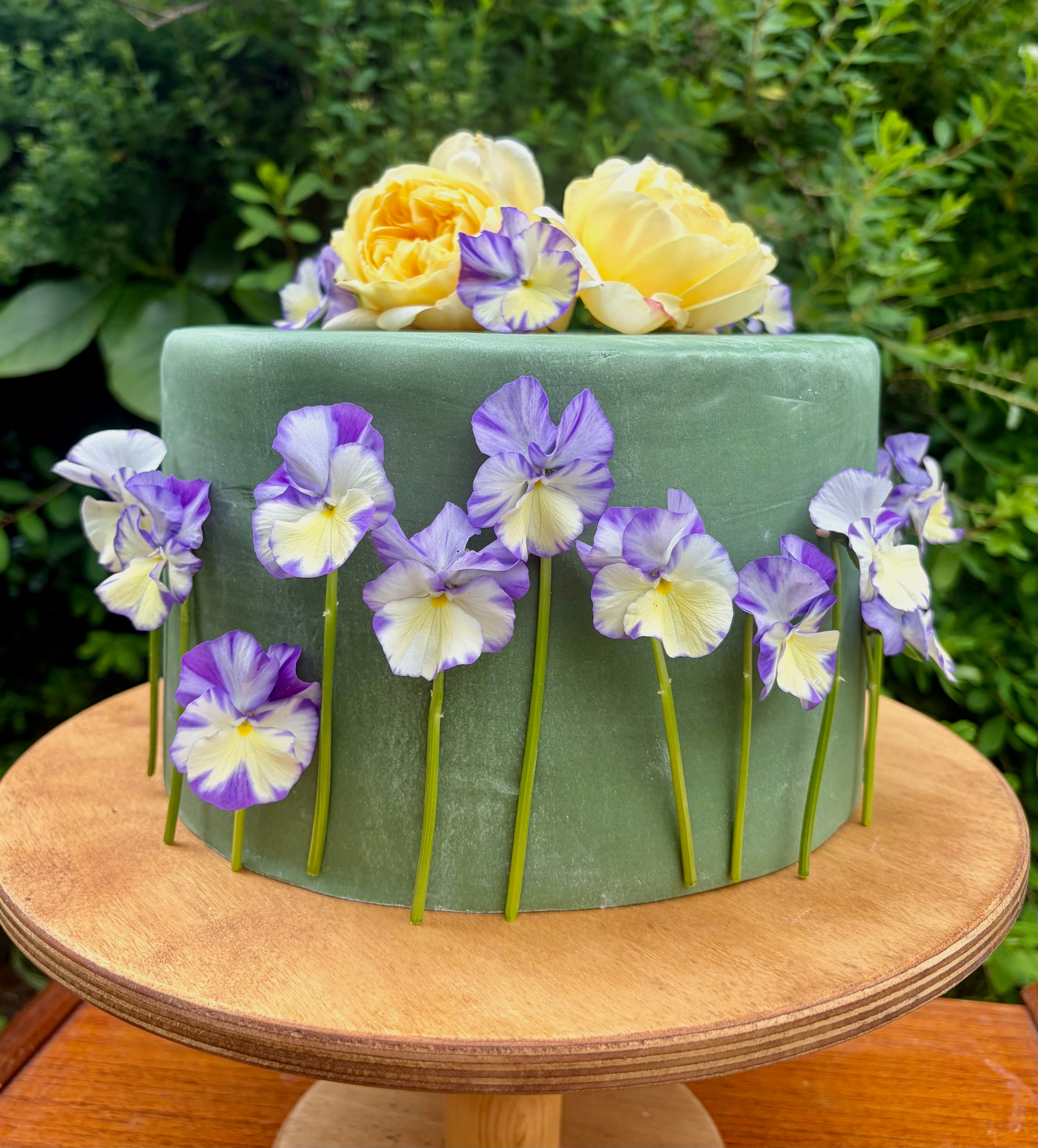 Green cake decorated with yellow and purple edible viola and roses on a wooden stand with a greenery background