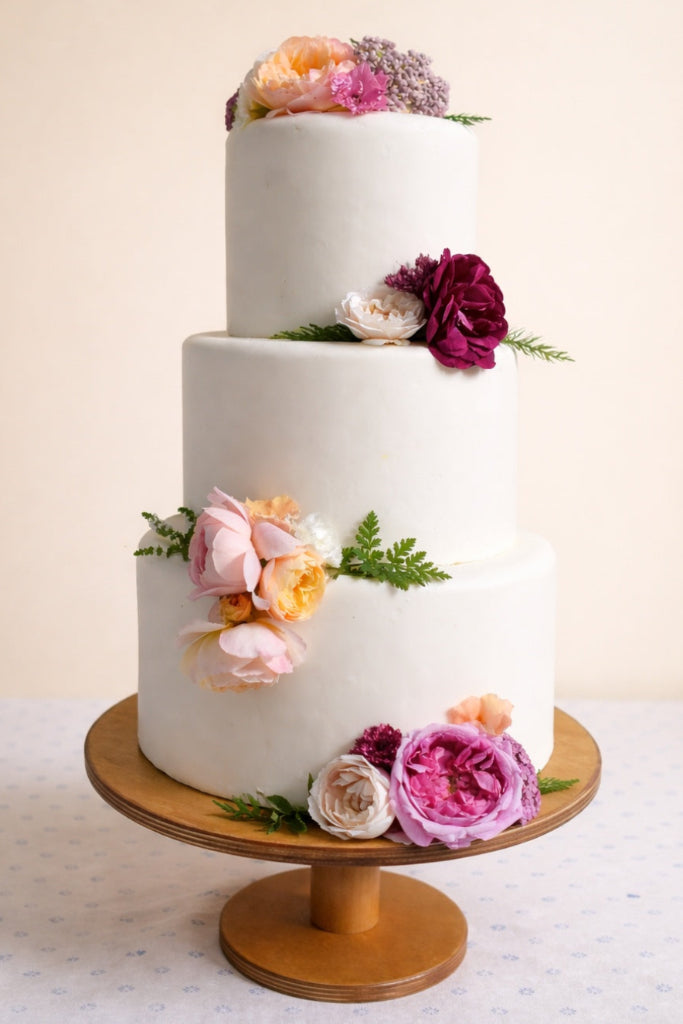 Three-tiered white wedding cake with edible flower decoration on a wooden stand against a light background
